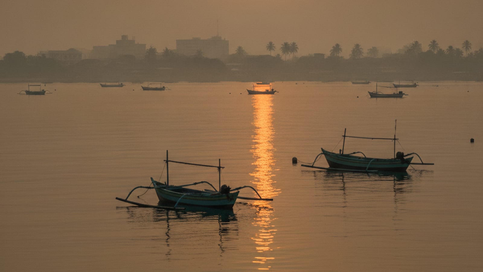 Hazy view over Dili harbour with traditional fishing boats in the foreground and the city skyline in the distance under an orange sky.