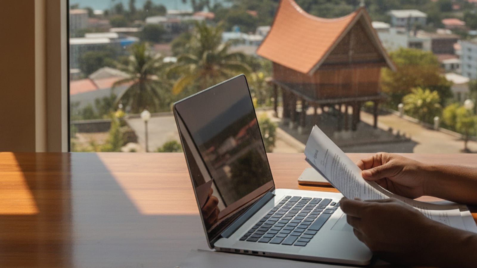 A person reviews tax documents and financial statements on a laptop, with a blurred background showing the city of Dili, Timor-Leste.